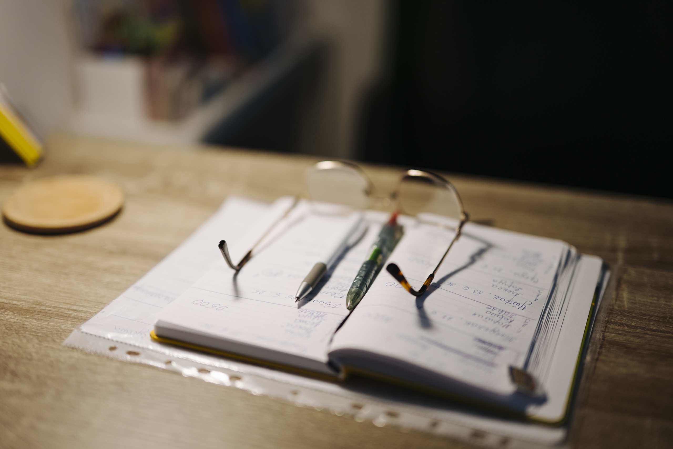 reading glasses and two pens resting on an open handwritten notebook on a warm wooden desk